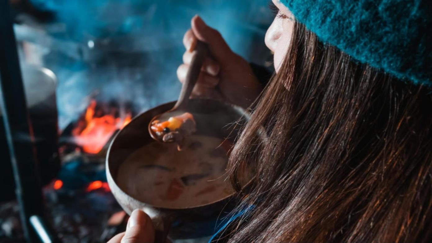 A girl eating soup from a wooden cup by an open fire
